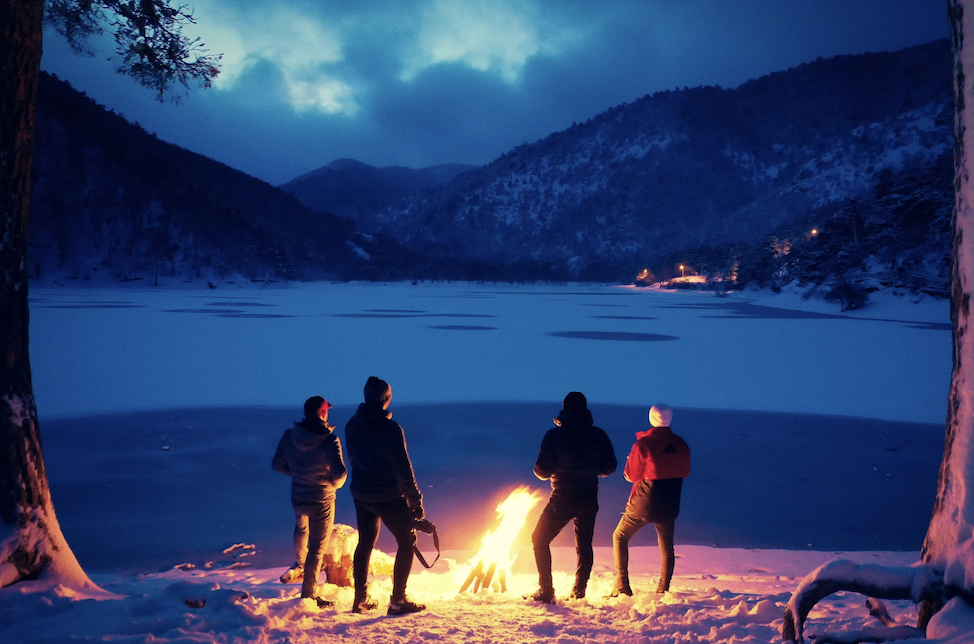 four people at a campfire near a lake in the snow looking at the mountains at night
